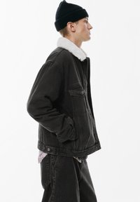 Giacca di jeans nera con colletto in pelliccia sintetica bianca, dotata di chiusure a pulsante e tasche laterali, abbinata a pantaloni di jeans scuri.