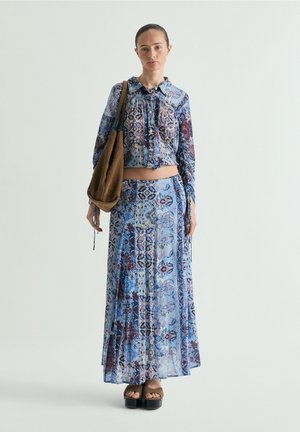 Frau steht, trägt ein blau gemustertes langärmeliges Croptop und passenden Maxirock, hält eine braune Schultertasche und trägt Plateau-Sandalen.