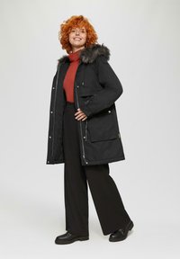 Schwarze Parkajacke mit Kunstfellkapuze, silbernen Beschlägen, Kordelzug und zwei Fronttaschen. Darunter ein roter Rollkragenpullover und schwarze Hose.