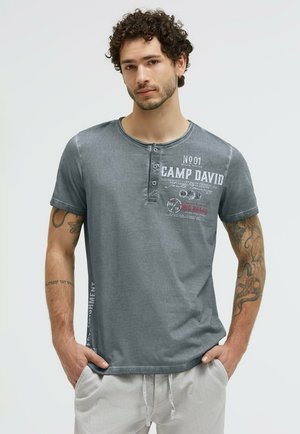 Mann mit lockigem Haar und Tattoos an beiden Armen trägt ein graues Camp David Kurzarmhemd und hellfarbene Hosen mit den Händen in den Taschen.