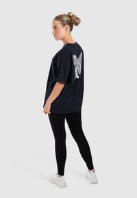 Schwarzes T-Shirt mit einem weißen Grafikdesign auf der Rückseite. Das Modell kombiniert es mit schwarzen Leggings und weißen Sneakers. Einfaches, lässiges Design.