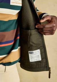 Zielona kurtka z miękkim polarowym wykończeniem i wewnętrzną kieszenią z białą metką. Posiada solidny zamek błyskawiczny oraz paskowaną bawełnianą koszulę pod spodem.