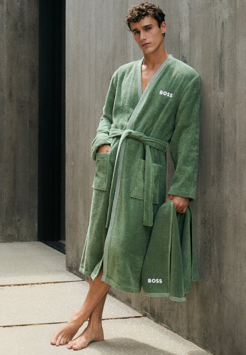 Jeune homme en peignoir vert BOSS avec une serviette assortie, debout pieds nus contre un mur en béton dans un cadre minimaliste.