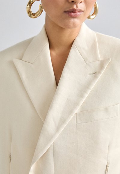 Blazer en lin couleur crème avec un large col, une structure d'épaule subtile et une poche avant avec un détail de broche. Grandes boucles d'oreilles en or.