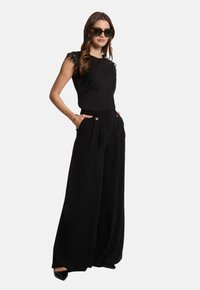 Zwarte mouwloze top met bloemenkant details, gecombineerd met zwarte wijde broek met voorplooien en zijzakken, aangevuld met hakken.