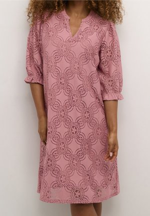 Femme portant une robe en dentelle rose poudré longueur genou avec motifs floraux et manches bouffantes trois-quarts.