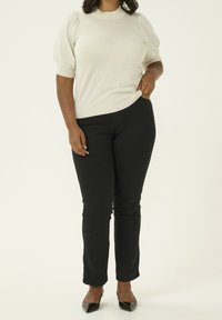 Vrouw in een crèmekleurige top met pofmouwen, zwarte slim fit jeans en zwarte pumps met puntige neuzen, staande voor een effen lichte achtergrond.