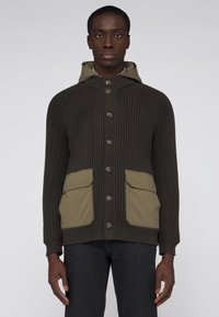 Mönstrad tröja i mörkbrun färg med knappslå framtill, huva och olivgröna fickdetaljer. Har ribbstruktur och raglanärmar.