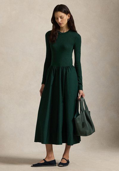 Mörkgrön klänning med långärmad, figursydd överdel och markerad midja, kombinerad med en grön läderväska och marinblå ballerinas med kontrasterande kant.