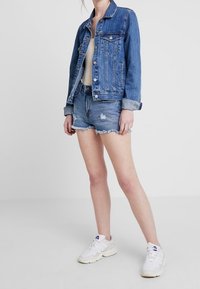 Chaqueta de denim en azul lavado medio con bolsillos frontales, combinada con pantalones cortos de denim deshilachados y zapatillas blancas con suelas texturizadas.