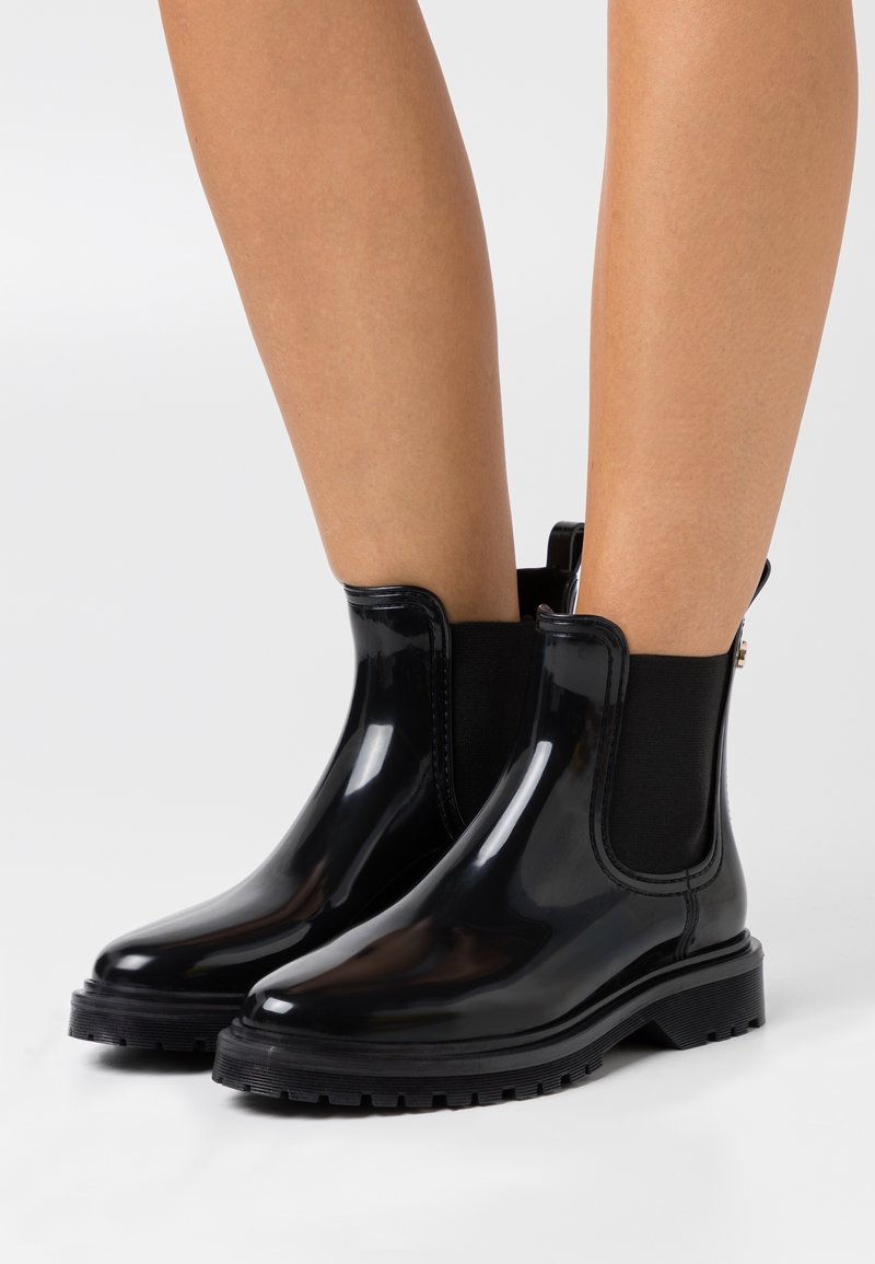 Botas de tobillo impermeables negras con un acabado brillante, paneles laterales elásticos y una suela resistente con un leve tacón.