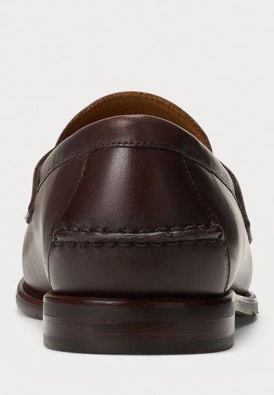 Zapato de piel marrón oscuro tipo mocasín visto desde atrás, mostrando los detalles de costura y el tacón de madera apilada sobre un fondo liso.