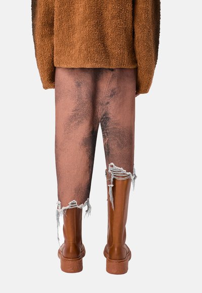 Brązowy sweter z polaru, wyblakłe różowe jeansy z przetarciami i postrzępionymi mankietami oraz brązowe botki za kostkę mają teksturowane powierzchnie i masywny design.