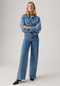 Vrouw met lang blond haar, gekleed in een blauwe spijkerjacket en hoge brede jeans, staand met gekruiste armen en zwarte hakken.