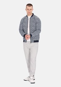 Giacca bomber navy con un motivo maculato, polsini a coste, abbinata a pantaloni della tuta grigi chiari e sneakers bianche, creando un look informale.