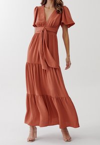 Maxiklänning i rostorange, med v-ringning, puffärmar, knytmidja och fackade volanger. Tillverkad av strukturerad tyg, lätt vikt.