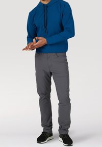 Mann trägt einen blauen Kapuzenpullover, graue Hose und schwarze Turnschuhe und steht mit vor dem Körper verschränkten Händen vor einem einfarbigen Hintergrund.