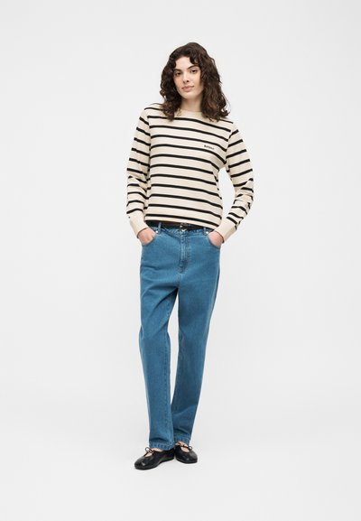 Femme aux cheveux bouclés portant une chemise à manches longues crème et noire rayée, un jean bleu et des ballerines noires, debout les mains dans les poches.