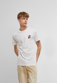 Balta medvilnė t-shirt su trumpomis rankovėmis, su mažos raudonos ir juodos rožės grafika kairėje krūtinės pusėje. Kasdienė figūra.