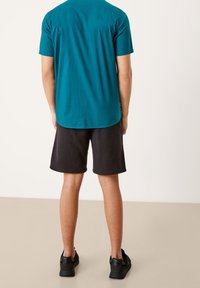 Maglietta a maniche corte teal con finitura testurizzata, abbinata a pantaloni corti neri. Scarpe da ginnastica nere completano l'outfit visto da dietro.