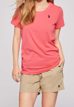 Kvinde iført koralrosa kortærmet t-shirt og beige shorts med sorte heste- og rytterlogoer, stående mod en ensfarvet baggrund.