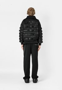 Chaqueta de bombardero negra con acentos de cinta en capas, que presenta un cuello, bolsillo trasero, y combinada con pantalones negros de corte holgado y calzado voluminoso.