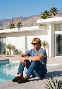 Homme portant des lunettes de soleil et des vêtements décontractés assis au bord d'une piscine devant une maison blanche moderne avec des palmiers et des montagnes en arrière-plan.