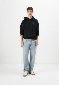 Un jeune homme se tenant debout, portant un sweat à capuche noir, un jean bleu clair et des baskets blanches, devant un fond blanc uni.