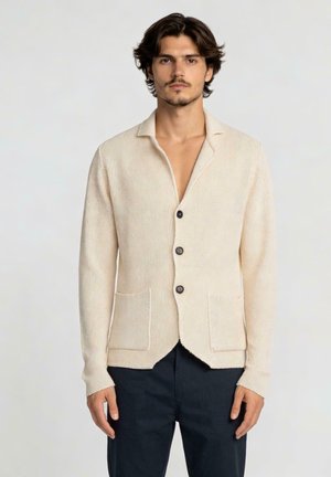 Homme portant un cardigan crème texturé boutonné avec deux poches avant et un pantalon foncé, debout devant un fond clair uni.