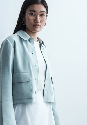 Νεαρή γυναίκα με μακριά σκούρα μαλλιά που φοράει διάφανα γυαλιά, χρυσούς κρίκους στα αυτιά, ανοιχτό μπλε σακάκι με κουμπιά και λευκό τοπ.