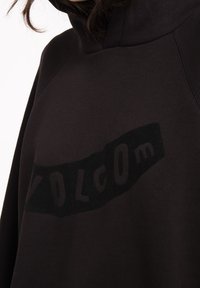 Musta huppari korkealla kaula-aukolla. Edessä on suuri, tumma "VOLCOM" -grafiikka, joka on valmistettu tekstuurimateriaalista pehmeällä pinnalla.