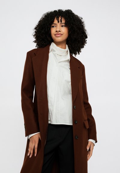 Rjav volnen plašč z dvorednim zapenjanjem, zavihani ovratniki in šestimi gumbi. Nošen prek bele majice in hlač z ozkimi črtami.