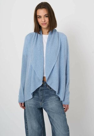 Młoda kobieta ubrana w jasnoniebieski otwarty kardigan, biały top i luźne niebieskie dżinsy, stojąca na jednolitym tle.