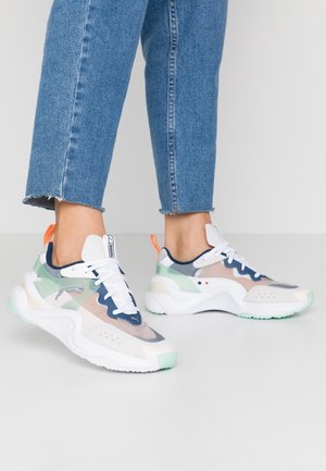 Person iført blå jeans og hvide multifarvede Puma sneakers står på en lys gulv mod en ensfarvet baggrund.