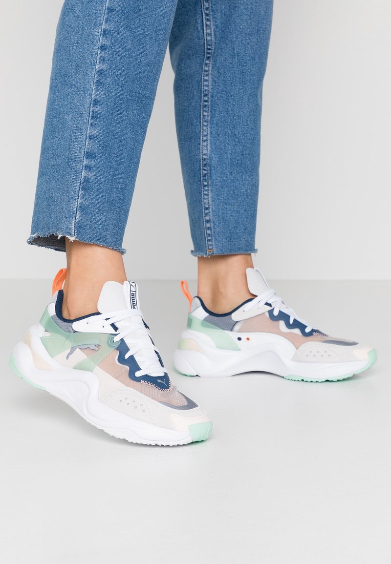 Personne portant un jean bleu et des baskets Puma blanches multicolores, debout sur un sol clair contre un fond uni.