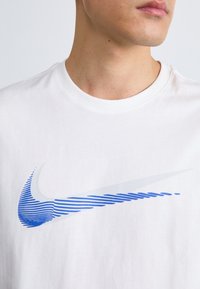 White cotton t-shirt featuring a blue and silver swoosh logo, with horizontal stripes on the logo and a round neckline.