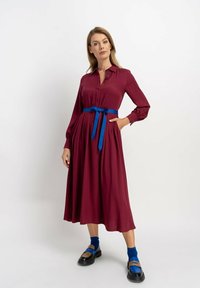 Burgundowa sukienka midi z długimi rękawami i zapięciem na guziki, podkreślona niebieskim paskiem; modelka nosi czarne buty i niebieskie skarpetki.