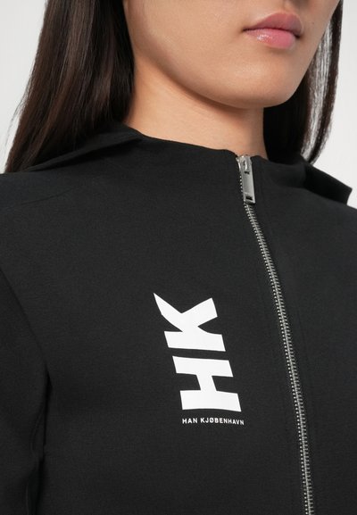 Pogled od blizu na osobu koja nosi crnu jaknu s patentnim zatvaračem s bijelim logotipom "HK HAN KJØBENHAVN" na prsima.