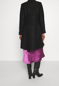 Abrigo negro de mezcla de lana con corte entallado, mangas largas y detalles de botones. Llevado sobre una falda de satén púrpura brillante y botas altas negras hasta la rodilla.