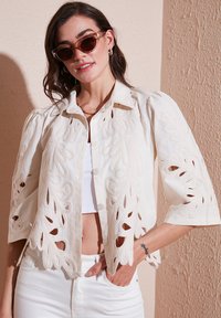 Blusa blanca con patrones florales bordados y recortados en las mangas, con cierre de botones en la parte delantera y mangas cortas con volumen. Usada sobre una blusa blanca.