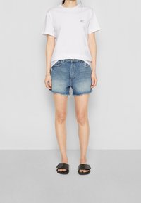 Žena v bílém tričku s nápisem "Crazy Sexy Cool", v potrhaných denimových šortkách a černých sandálech na plocho, stojí na šedé podlaze.