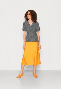 Mulher a usar óculos de sol laranja, uma blusa preta de manga curta com padrão floral, uma saia midi amarelo-mostarda e sapatos de salto abertos laranja, de pé contra um fundo simples.