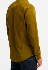 Sinapinkeltainen nappipaita, jossa on klassinen kaulus, pitkät hihat ja sileä pinta, yhdistetty tummiin farkkuihin.