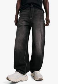 Blugi negri cu croială largă și cusături gri deschis. Realizați din denim, cu închidere frontală cu nasturi și fermoar. Purtați cu adidași albi.