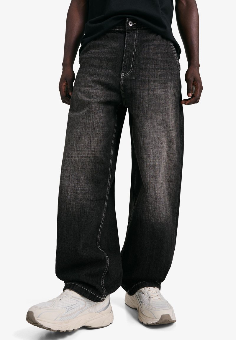 Črne kavbojke širokih nog z svetlo sivim šivom. Iz džinsa, s sprednjim zapiranjem na gumb in zadrgo. Nošene z belimi supergami.