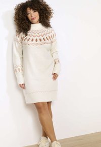Pletené krémové šaty z svetrového materiálu s geometrickými vzory v světle hnědé a korálové barvě. Mají vysoký límec a žebrované manžety. Hladká textura.