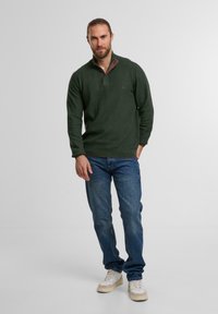 Mann mit zurückgebundenen Haaren, der einen dunkelgrünen Pullover, blaue Jeans und weiße Sneakers trägt und vor einem hellen, einfarbigen Hintergrund steht.