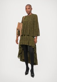 Robe à manches longues en vert foncé avec un motif subtil, dotée de volants à l'ourlet et associée à des collants noirs et des chaussures à bouts pointus.