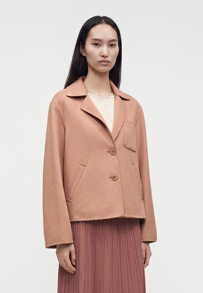 Femme aux longs cheveux foncés portant une veste marron clair boutonnée sur un haut crème et une jupe plissée rose poudré sur un fond uni.