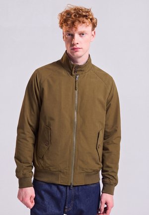 Junger Mann mit lockigem roten Haar, der eine olivgrüne Jacke mit Reißverschluss und blaue Jeans trägt, steht vor einem schlichten hellen Hintergrund.